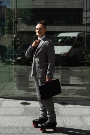 Lächelnder Geschäftsmann in Anzug und Rollschuhen, der Krawatte fixiert und in die Kamera an der Glasfassade blickt