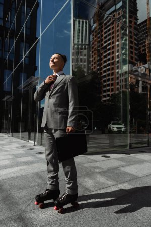 Geschäftsmann in grauem Anzug und Rollschuhen mit Aktentasche und Krawatte auf einer städtischen Straße in der Nähe eines modernen Gebäudes