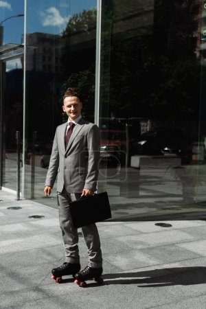 full length of smiling businessman in roller skates standing with black briefcase near glass facade of modern building