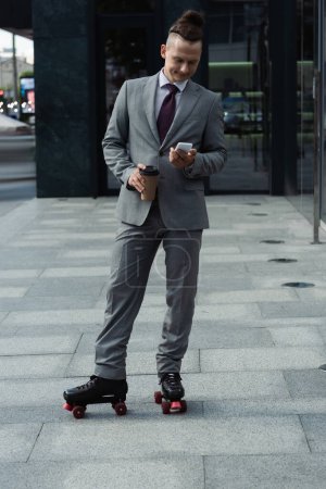 stylischer Geschäftsmann in formeller Kleidung und Rollschuhen mit Smartphone und Imbissgetränk auf der Stadtstraße
