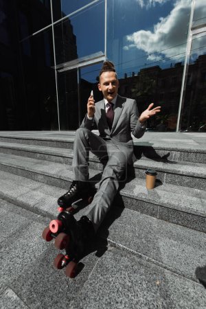 Foto de Feliz hombre de negocios en patines de ruedas sosteniendo el teléfono inteligente y apuntando con la mano mientras está sentado cerca del café para salir al aire libre - Imagen libre de derechos
