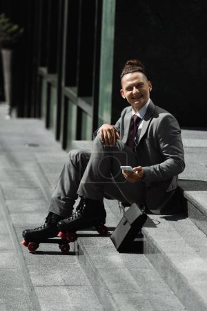 Photo for Cheerful businessman in roller skates and wireless earphones holding cellphone and looking at camera while sitting on stairs outdoors - Royalty Free Image