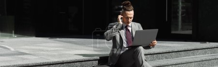Photo for Serious businessman with laptop adjusting earphone while sitting on stairs on street, banner - Royalty Free Image
