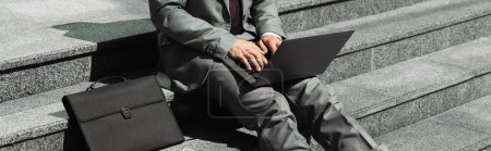 Teilaufnahme eines Geschäftsmannes im grauen Anzug, der auf einer Treppe neben einer schwarzen Aktentasche sitzt und auf Laptop tippt, Banner