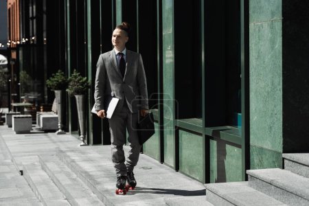 Photo for Happy man in business suit and roller skates holding laptop and briefcase while skating on city street - Royalty Free Image