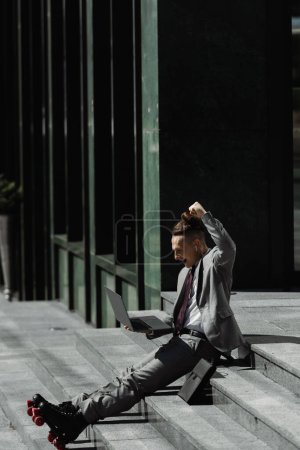 Photo pour Homme d'affaires excité en patins à roulettes montrant geste de victoire tout en étant assis sur les escaliers de la rue de la ville - image libre de droit
