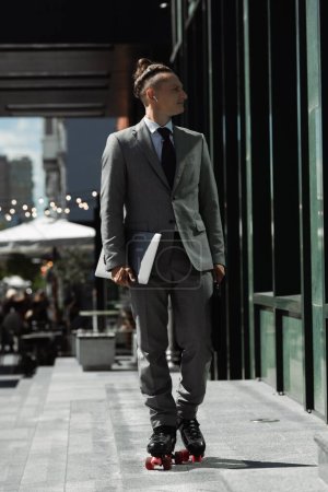 junger Geschäftsmann im grauen Anzug mit Laptop beim Rollschuhlaufen auf der Stadtstraße