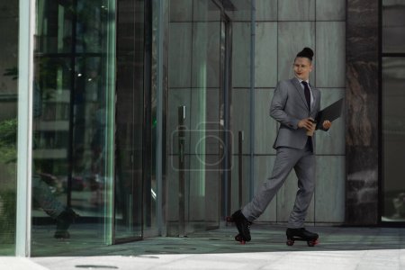 jeune homme d'affaires en patins à roulettes et costume gris tenant café pour aller et ordinateur portable près du bâtiment avec portes en verre
