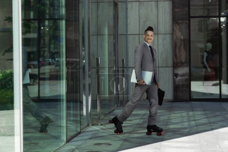 businessman in roller skates holding laptop and briefcase while smiling at camera near building with glass doors