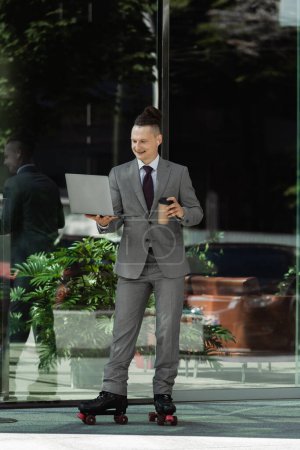 smiling man in grey suit and roller skates holding coffee to go and looking at laptop near green plant behind glass facade