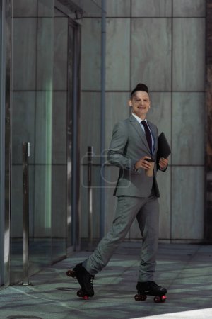 Foto de Sonriente hombre de negocios en patines de ruedas sosteniendo portátil y taza de papel mientras patina fuera del edificio con puertas de vidrio - Imagen libre de derechos