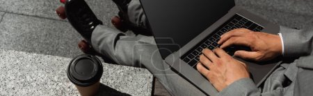 Photo for Top view of businessman in roller skates and suit typing on laptop near paper cup on urban street, banner - Royalty Free Image