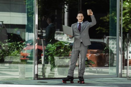 Photo for Smiling businessman in roller skates standing with laptop and coffee to go in raised hand near glass building - Royalty Free Image