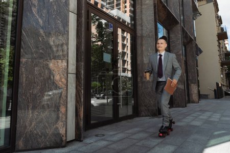 smiling man in suit and roller skates riding with coffee to go along building on urban street