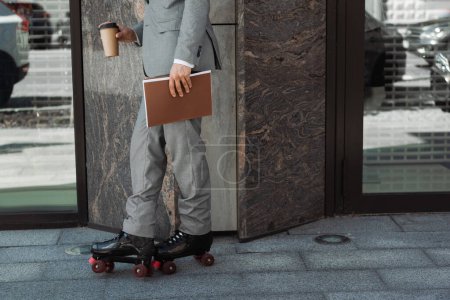 Foto de Vista recortada del patinador en traje de pie con bebida para llevar y carpeta cerca del edificio en la ciudad - Imagen libre de derechos