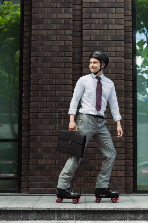 young businessman with briefcase roller skating on urban street and looking back