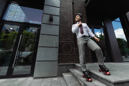 full length of businessman in roller skates and formal wear fixing tie and looking away near contemporary building