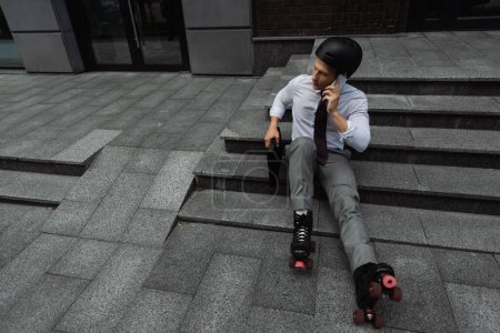 Blick aus der Vogelperspektive auf Geschäftsmann mit Helm und Rollschuhen, der auf dem Smartphone spricht, während er im Freien auf der Treppe sitzt