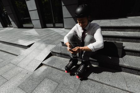 high angle view of businessman in roller skates sitting on stairs and messaging on smartphone