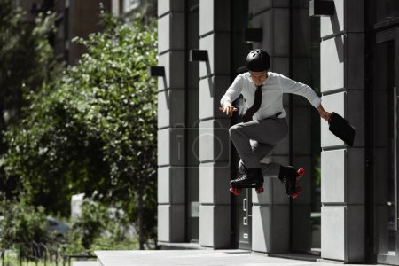 Foto de Longitud completa de hombre de negocios en patines de ruedas y el casco saltando mientras hace truco en la calle cerca del edificio - Imagen libre de derechos