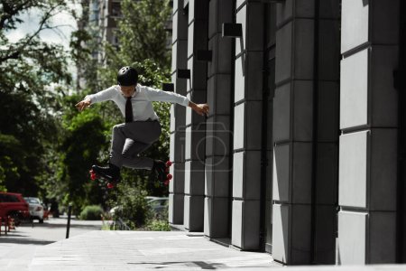 Foto de Longitud completa de hombre de negocios en casco y patines saltando mientras hace truco en la calle de la ciudad - Imagen libre de derechos