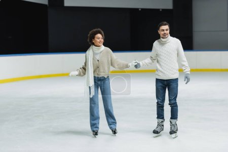Smiling multiethnic couple in sweaters and jeans skating during date on ice rink 