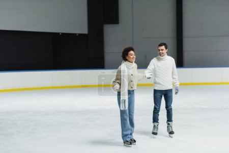 Foto de Positivo interracial pareja patinaje sobre hielo y de la mano durante la fecha en la pista de patinaje - Imagen libre de derechos
