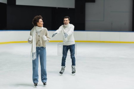 Téléchargez les photos : Homme souriant étendant les mains à la petite amie afro-américaine patinoire sur la patinoire - en image libre de droit