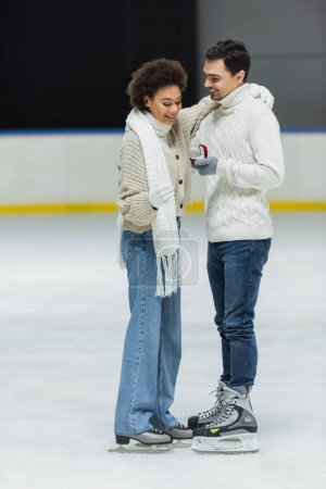 Sonriente mujer afroamericana mirando anillo de propuesta cerca de novio en pista de hielo 