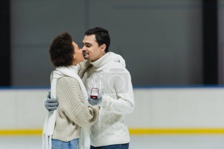 Foto de Joven sosteniendo anillo de propuesta y besando novia afroamericana en pista de hielo - Imagen libre de derechos