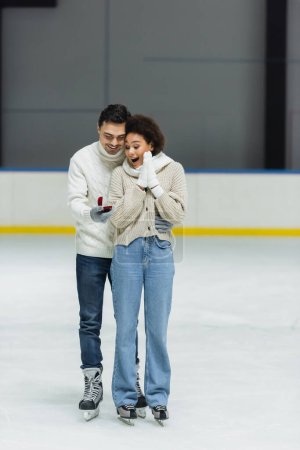 Photo pour Un homme souriant tenant une bague de fiançailles près d'une copine afro-américaine excitée sur une patinoire - image libre de droit