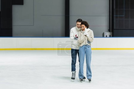 Man holding proposal ring and hugging african american girlfriend on ice rink 