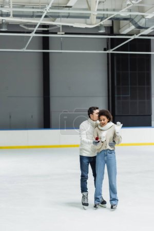 Man hugging african american girlfriend and holding engagement ring on ice rink 