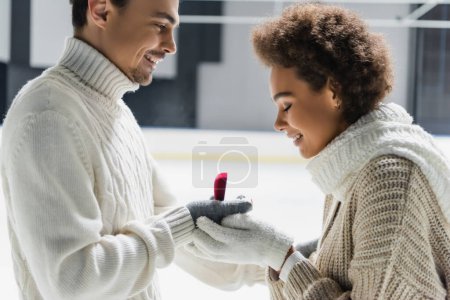 Foto de Vista lateral del hombre en suéter sosteniendo anillo de compromiso cerca de novia afroamericana en pista de hielo - Imagen libre de derechos