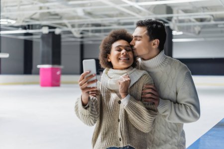 Téléchargez les photos : Jeune homme embrassant petite amie afro-américaine prenant selfie sur la patinoire - en image libre de droit