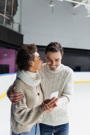 Photo for Smiling interracial couple hugging and using smartphone on ice rink - Royalty Free Image