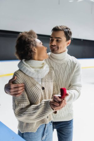 Téléchargez les photos : Sourire femme afro-américaine regardant petit ami avec bague de demande en mariage floue sur la patinoire - en image libre de droit