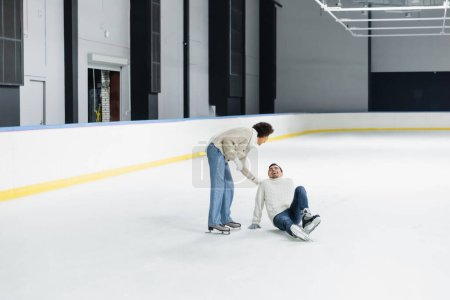 African american woman standing near cheerful boyfriend sitting on ice rink 