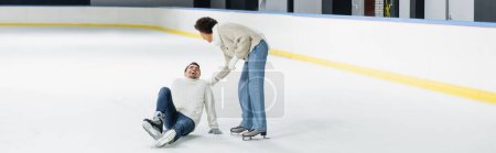 Mujer afroamericana joven ayudando novio sentado en pista de hielo, pancarta 