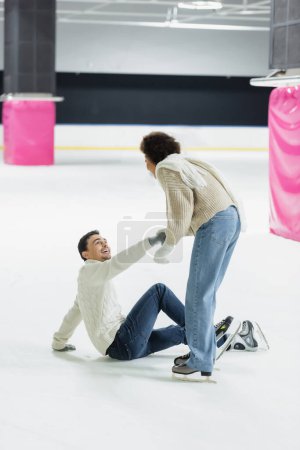 African american woman in sweater helping boyfriend on ice rink 