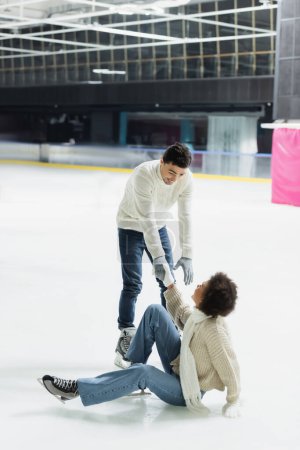 Young man giving hand to african american girlfriend falling on ice rink 