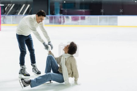 Smiling man giving hand to african american girlfriend on ice rink 