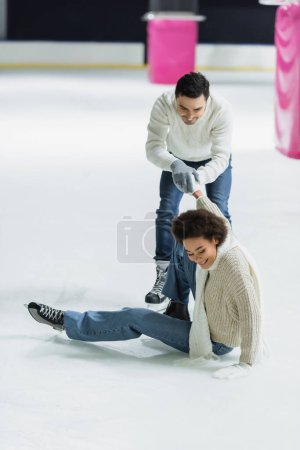 Smiling african american woman sitting on ice near boyfriend on rink 