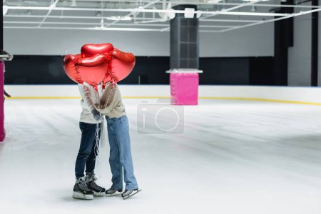 Couple hugging and holding red balloons in shape of heart on ice rink