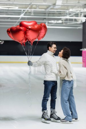 Téléchargez les photos : Vue latérale d'un homme souriant tenant des ballons en forme de cœur près d'une petite amie afro-américaine sur une patinoire - en image libre de droit