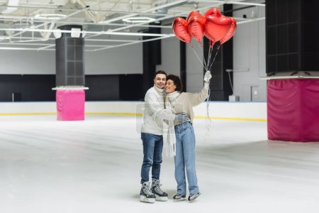 Positive interracial couple hugging and holding balloons in shape of heart on ice rink 
