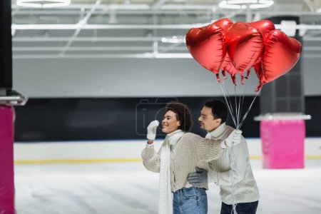 Young man hugging cheerful african american girlfriend in sweater holding red heart shaped balloons on ice rink 