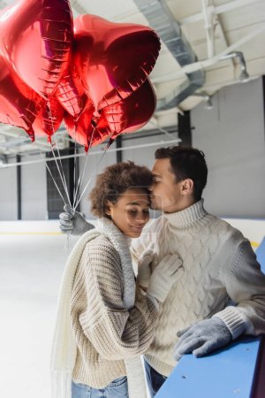 Junger Mann in Handschuhen und Pullover küsst afrikanisch-amerikanische Freundin und hält herzförmige Luftballons auf Eisbahn 
