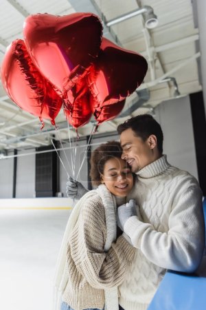 Young man in sweater hugging african american girlfriend and holding red heart shaped balloons on ice rink 