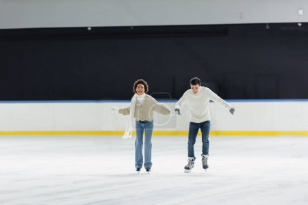 Téléchargez les photos : Couple multiethnique souriant en chandails tenant la main pendant le patinage sur glace sur la patinoire - en image libre de droit